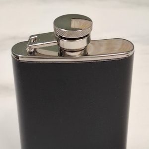 May include: A rectangular pocket flask featuring a smooth black exterior finish. The top section, including the screw-on cap and the hinged attachment, is constructed from polished silver-tone metal with a textured grip on the cap edge.