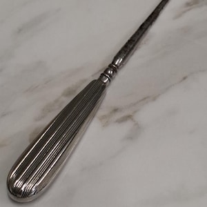 May include: A silver shoe horn with a long, slender handle and a curved hook. The handle features a ribbed design. The item has a vintage appearance and is likely made of metal.
