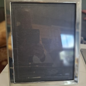 Puede incluir: Un marco de fotos rectangular plateado con un borde de cuentas. El marco tiene un interior gris oscuro y está diseñado para contener una fotografía. El marco es de metal y tiene una superficie reflectante.