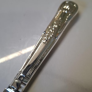 May include: Close-up of a silver knife with ornate floral detailing on the handle. The knife blade is visible at the bottom, and the handle features a decorative pattern. The knife is likely part of a silverware set.