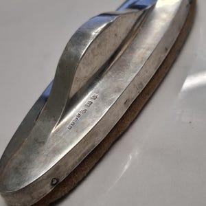 May include: Silver-plated oval-shaped object with a brown suede-like surface and a handle. The object has the inscription "BIRMINGHAM 1919" on the side.