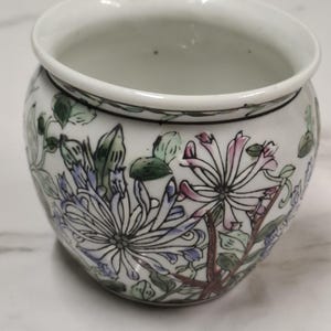 Pode incluir: Um pequeno vaso de cerâmica branca de formato arredondado com um design floral pintado à mão. O exterior apresenta ilustrações detalhadas de crisântemos roxos e rosas com folhagem verde, delineados com linhas pretas finas.