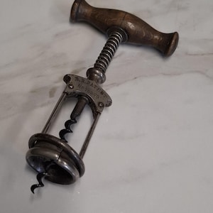 Könnte beinhalten: Ein antiker Metallkorkenzieher mit einem dunkelbraunen, T-förmigen Holzgriff. Der Metallrahmen verfügt über einen Spiralfedermechanismus und eine zentrale Metallplatte mit der Gravur D.R. PATENT No 70879. Eine scharfe, spiralförmige Metallspindel ragt hervor.