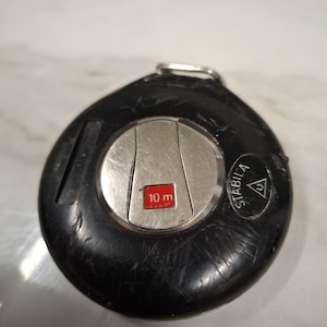 Op de afbeelding: Een zwart en zilver meetlint met een 10 m meetindicator. De tape heeft een cirkelvormig ontwerp met de merknaam "STABILA" zichtbaar. De rolmaat is een hulpmiddel voor de bouw en andere meetbehoeften.