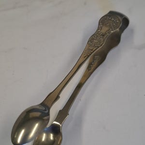 Könnte beinhalten: Silberfarbene Zuckerklemme mit aufwendigen floralen und scrollartigen Details. Die Zange hat zwei löffelförmige Enden zum Schöpfen von Zucker. Der Griff weist dekorative Gravuren auf. Ein Vintage-Küchenutensil.
