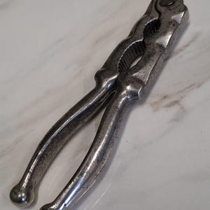 May include: A vintage-style metal nutcracker featuring a weathered, dark silver-toned finish. The tool has two long, curved handles with rounded ends and a central, serrated gripping section designed for cracking shells, showing signs of age.
