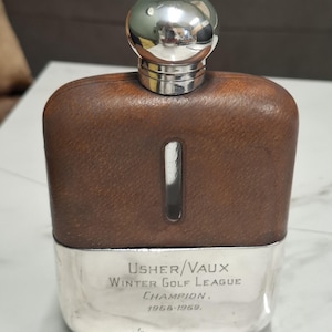 Puede incluir: Un frasco vintage con cuerpo de cuero marrón y base y tapón plateados. La base está grabada con "USHER/VAUX Winter Golf League Champion, 1958-1959."