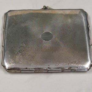 May include: A rectangular silver-toned case with a hinged lid and a decorative, swirling pattern on the surface. The edges are scalloped, and a small clasp secures the lid. The case has a small, oval-shaped accent in the center.