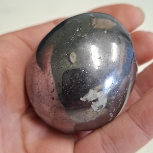 May include: A smooth, polished, rounded stone with a metallic, dark gray, reflective surface. The stone features irregular patches of light gray and white mineral inclusions embedded within the dark, glossy, and slightly uneven exterior texture.