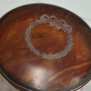 Puede incluir: Un recipiente de metal plateado con tapa cobriza. La tapa presenta un diseño de corona grabado con un lazo en la parte superior. El recipiente tiene una superficie brillante y reflectante.