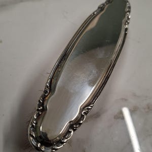 Op de afbeelding: Een ovale haarborstel in zilverkleur met een gepolijste, reflecterende metalen achterkant voorzien van een sierlijke, in reliëf gemaakte rand. De borstel heeft donkere, fijne haren die zichtbaar zijn langs de randen van het decoratieve frame.