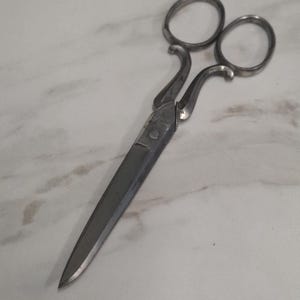 Puede incluir: Un par de tijeras de metal antiguas con mangos curvos y orificios para los dedos. Las hojas son afiladas y puntiagudas, y las tijeras están hechas de un metal de color plateado. Es probable que las tijeras se utilicen para cortar tela o papel.