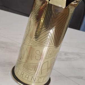 May include: A brass vase with a flared, petal-like top and a cylindrical body. The vase is engraved with geometric patterns and the word "FRANCE". The vase has a circular base and a polished finish.