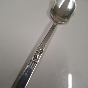 Peut inclure: Une cuillère en métal argenté dotée d'un cuilleron de forme carrée et d'un manche plat. Le manche est orné d'une série de lignes horizontales gravées et d'un motif floral central en relief, avec une finition métallique polie et brillante.
