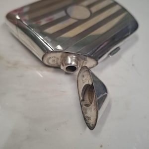 Puede incluir: Una petaca rectangular plateada con tapa abisagrada y un pico curvo separado. La superficie de la petaca presenta un patrón de rayas, y el pico está separado. La petaca es probablemente de metal.