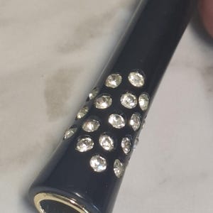 May include: A cylindrical black cigarette holder featuring a cluster of clear, sparkling rhinestones embedded on the exterior surface. The tip of the holder is finished with a smooth, metallic gold-toned rim, providing a contrast to the dark body.