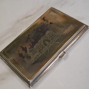 May include: A rectangular, silver-toned metal cigarette case. The case features an etched design on the lid, with a detailed pattern and a small clasp. The surface shows signs of age and wear, adding to its vintage appeal.