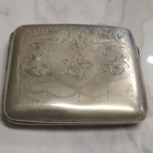 Könnte beinhalten: Ein rechteckiges Etui aus silberfarbenem Metall mit einem komplizierten, gravierten Rankenmuster auf der Oberseite. In der Mitte des Deckels befindet sich eine dekorative Schildform mit einem verzierten, stilisierten Monogramm der Buchstaben JS.