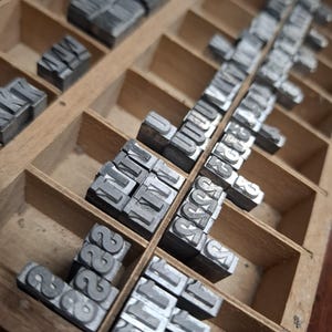 May include: A wooden letterpress type case containing numerous individual metal letter blocks arranged in grid compartments. Each silver-colored block features a raised character on its top surface, organized for manual printing.