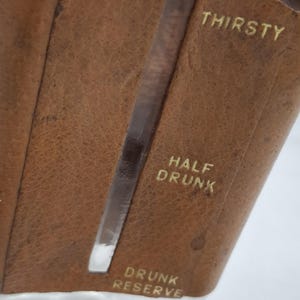 Könnte beinhalten: Eine Karaffe aus klarem Glas, umhüllt von einer braunen Lederhülle mit einem vertikalen Ausschnitt zur Anzeige des Flüssigkeitsstands. Das Leder ist mit dem Text THIRSTY oben, HALF DRUNK in der Mitte und DRUNK RESERVE unten geprägt.