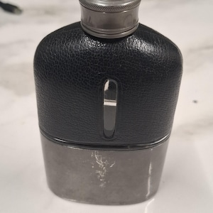 Peut inclure: Une flasque de poche avec une partie supérieure noire texturée et une base métallique lisse. Elle est dotée d'un bouchon à vis argenté avec un bord moleté et d'une fenêtre verticale étroite et réfléchissante centrée sur la section noire du récipient.