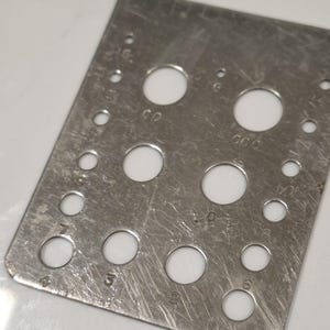 Könnte beinhalten: Ein rechteckiges, silberfarbenes Metallmessgerät mit mehreren kreisförmigen Löchern unterschiedlicher Größe. Das Messgerät hat eingravierte Zahlen neben den Löchern, die ihren Durchmesser angeben. Die Metalloberfläche weist sichtbare Kratzer auf.