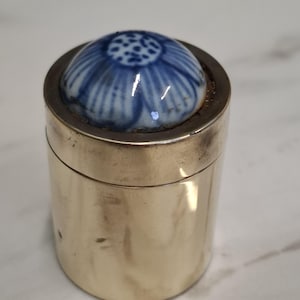 May include: A small, cylindrical container with a silver-coloured metal body. The lid has a blue and white floral design on a rounded, ceramic top. The container is likely for storing small items.