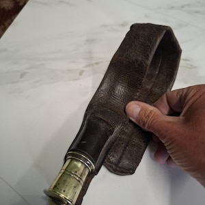 May include: A brown leather sheath with a brass and dark wood handle. The sheath is worn and textured, with a loop at the top. The handle has a decorative brass section and a rounded end.