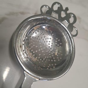 May include: A silver-coloured metal tea strainer featuring a circular bowl with a perforated base and a decorative, ornate handle attachment. The strainer has a polished, reflective surface and a series of small, evenly spaced holes for straining.