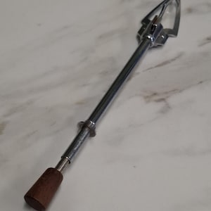 May include: A chrome-plated metal tool featuring a long, slender cylindrical shaft, a spring-loaded claw mechanism at one end, and a dark brown wooden handle at the opposite end. The device has a polished, reflective finish throughout the metal body.