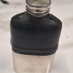 May include: A pocket flask featuring a silver-toned metal base and a black leather-wrapped upper section. The flask has a cylindrical silver-toned screw-top cap with a textured, ribbed edge. The body is rectangular with rounded corners and edges.