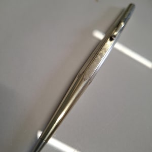 以下が含まれることがあります： 先細りのペン先と上部バレルにテクスチャ加工されたクロスハッチパターンが特徴の銀色の金属製ボールペン。このペンには、スリムな一体型ポケットクリップと、下部に滑らかで光沢のある仕上げが施されています。