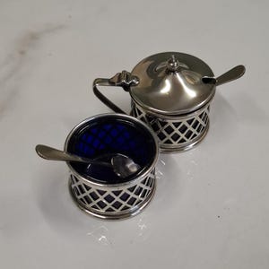 May include: Two silver-toned metal condiment holders with cobalt blue glass inserts. One holder features a hinged lid and a handle, while the other is open-topped. Both containers have a lattice-style metal overlay and include small metal spoons.
