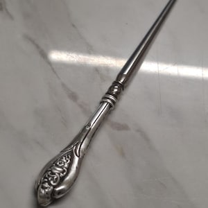 Può includere: Un tirabottone in metallo color argento con un manico decorato da motivi floreali in rilievo. L'utensile presenta un'asta lunga e sottile che termina con un piccolo gancio ricurvo, con fasce decorative a rilievo tra il manico e l'asta.