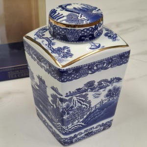 Könnte beinhalten: Eine quadratische Teedose aus Keramik mit blau-weißem Weidenmuster und Goldrand am Deckel sowie an den oberen Kanten. Das Design zeigt traditionelle Landschaftsszenen mit Pagoden, Bäumen und einer Brücke. Der Deckel ist rund und abnehmbar.