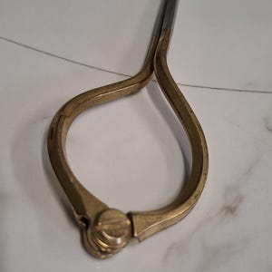 Könnte beinhalten: Ein Messzirkel aus Messing mit einem geschwungenen, tropfenförmigen Griff und einer zentralen Stellschraube. Das Werkzeug verfügt über zwei lange, schlanke Metallspitzen, die nach oben ragen, mit einer polierten Oberfläche und sichtbaren Gebrauchsspuren.
