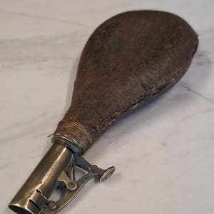 Könnte beinhalten: Eine Vintage-Pulverflasche aus braunem Leder mit einem Metallausguss und einem federbelasteten Mechanismus. Die Flasche hat eine bauchige Form mit einem Metallausguss und einem Klapphebel zum Ausgeben von Pulver. Das Design deutet darauf hin, dass sie zum Aufbewahren und Ausgeben von Schießpulver verwendet wurde.