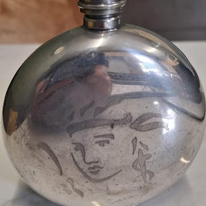 May include: A round, silver-coloured metal flask with a polished, reflective surface. The front features a simple, etched line drawing of a human face with stylised hair. It has a cylindrical, ribbed screw-top cap at the centre of the top edge.
