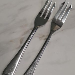May include: Two silver-toned metal cocktail forks with three tines, featuring a distinct pointed hook on the outermost tine of each fork. The handles have a smooth, tapered design with a rounded end, showcasing a polished, reflective finish.