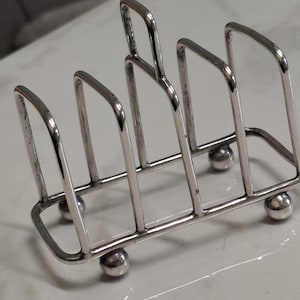 May include: A silver-colored metal toast rack with a rectangular base and four vertical dividers. The rack has a handle and rests on four small, spherical feet. The design is simple and functional.