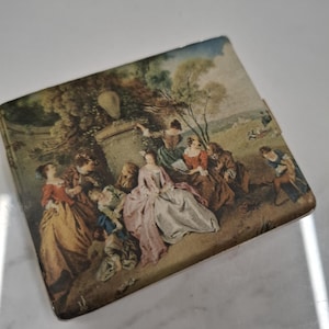 May include: A rectangular compact case featuring a printed scene of a group of people in 18th-century attire gathered outdoors near a stone pedestal and trees. The illustration displays figures in colourful dresses and coats in a park setting.