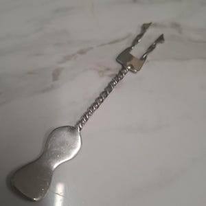 Peut inclure: Une fourchette à cocktail en métal argenté dotée d'une tête à deux dents, d'un manche fin à la texture torsadée rappelant une corde et d'une base plate, lisse et en forme de sablier. L'ustensile présente une finition métallique polie.
