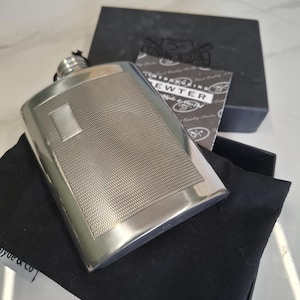 Peut inclure: Une flasque en métal argenté avec un panneau central texturé et une bordure lisse. La flasque est dotée d'un bouchon à vis et est présentée dans une boîte noire avec une carte portant l'inscription Introducing Pewter, Style & Quality.