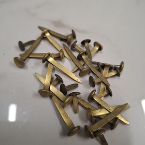 May include: A collection of brass-coloured metal paper fasteners, also known as brads, featuring a round head and two flexible prongs extending from the back. These fasteners are scattered in a cluster, showing their metallic finish and structure.