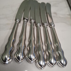 May include: A collection of six silver butter knives. Each knife features a curved blade and a handle with a wavy, decorative pattern. The knives are displayed in a row, reflecting light.