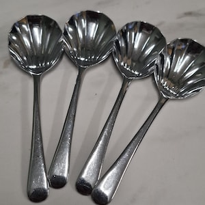 Può includere: Quattro cucchiai d'argento da portata con una coppetta a forma di conchiglia e manici lunghi e sottili. I cucchiai sono disposti in fila, riflettendo la luce. Probabilmente per servire salse o altre piccole porzioni.