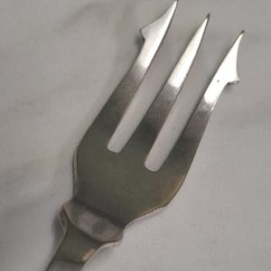 May include: A silver-toned metal seafood fork featuring three tines. The two outer tines are designed with a distinct, sharp barb or hook protruding from the side, while the centre tine is straight and pointed, all finished with a smooth, polished surface.