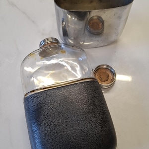 Könnte beinhalten: Eine Vintage-Flasche mit einem transparenten Glaskörper und einer schwarzen Lederhülle. Die Flasche hat einen silberfarbenen Metallaufsatz und einen passenden Verschluss. Eine ovale, silberfarbene Metalltasse befindet sich hinter der Flasche.