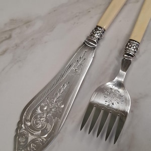 May include: A silver-plated fish knife and fork set featuring intricate floral engravings on the blades and tines. The handles are a smooth, off-white cream color, connected to the metal by ornate, textured silver-toned collars with floral details.