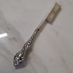 May include: An antique silver-handled brush with a floral design. The bristles are off-white and show signs of wear. This vintage grooming tool has a decorative handle.